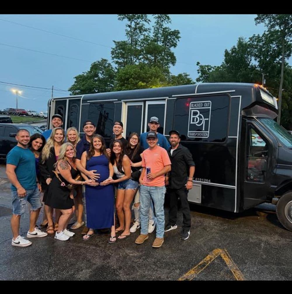 Group of friends celebrating a pregnant woman beside a black party bus at an outdoor event.