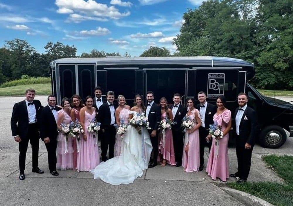 Wedding party in elegant attire posing together near a black party bus on a sunny day.
